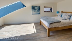 A bedroom with a sloped ceiling, a skylight, a bed with a wooden frame, a small window, and a painting on the wall. - The Wave (Bridport)