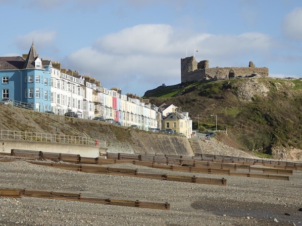 Un pueblo costero con casas de colores en terrazas, un castillo de piedra histórico sobre una colina y una playa de guijarros en primer plano.
