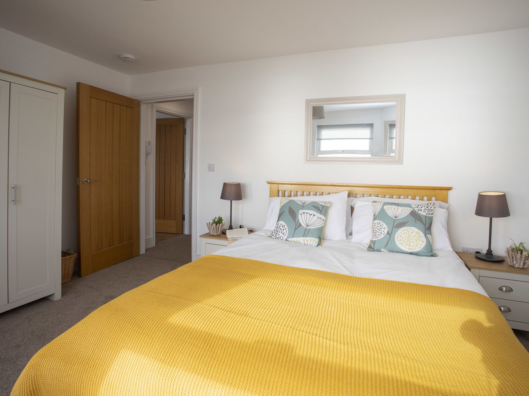 Un dormitorio con cama, lámparas de noche, mesita de noche con lámpara, espejo en la pared y una puerta que da a otra habitación.