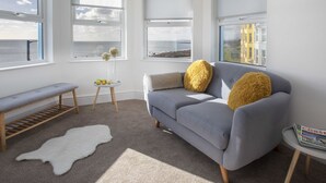 Een moderne woonkamer met een grijze bank, twee gele kussens, een witte bankje en een klein rond tafeltje met een tijdschrift.