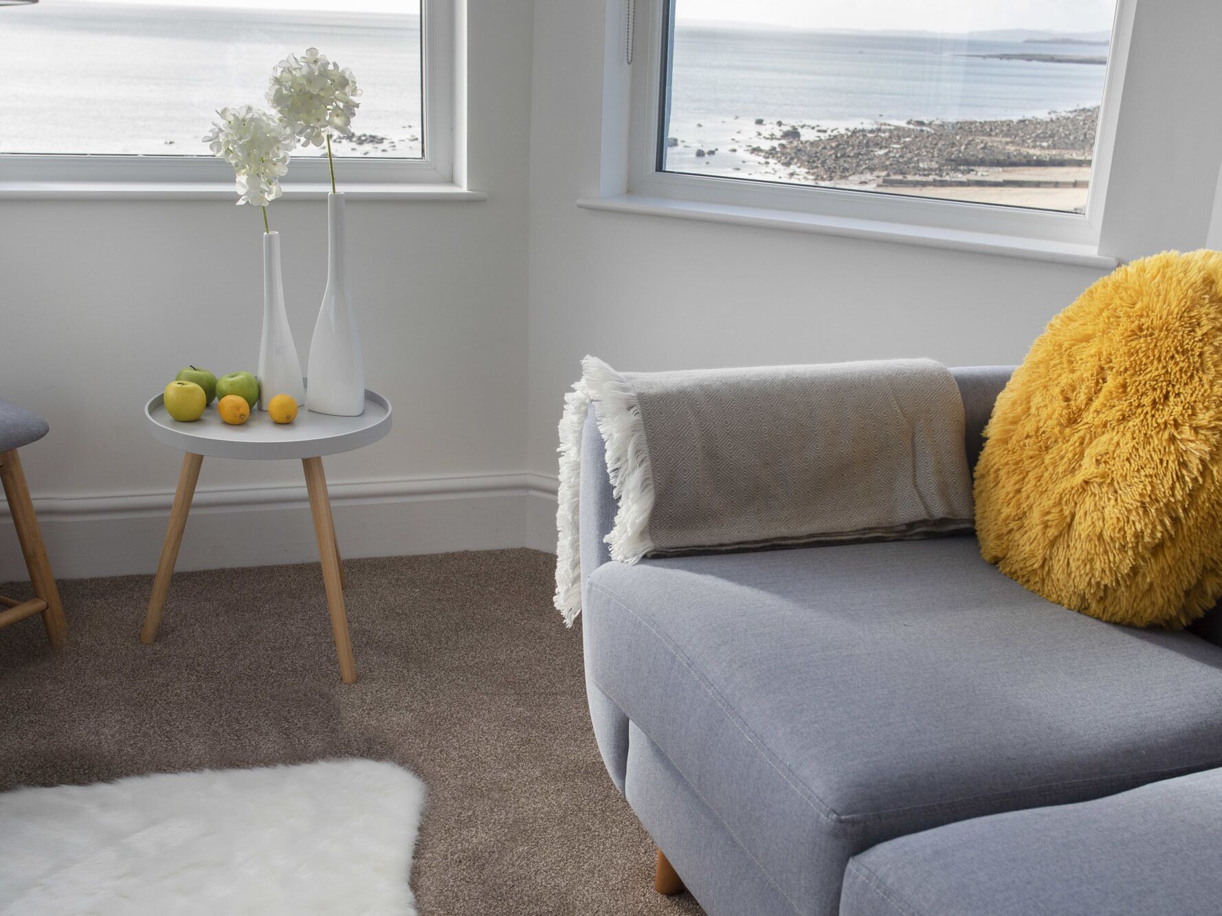 Una sala de estar con un sofá gris, un cojín amarillo y esponjoso, una pequeña mesa blanca con frutas y un florero, y vistas al mar a través de amplias ventanas.