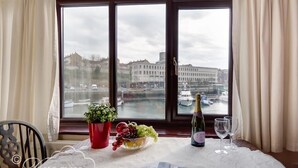 Una mesa de comedor con una botella de champán, una copa, un bol de fruta y un menú. La mesa está frente a una ventana con vistas a un paisaje urbano y una masa de agua.