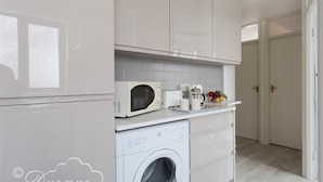 Una cocina compacta con armarios blancos, microondas, cafetera y lavadora.
