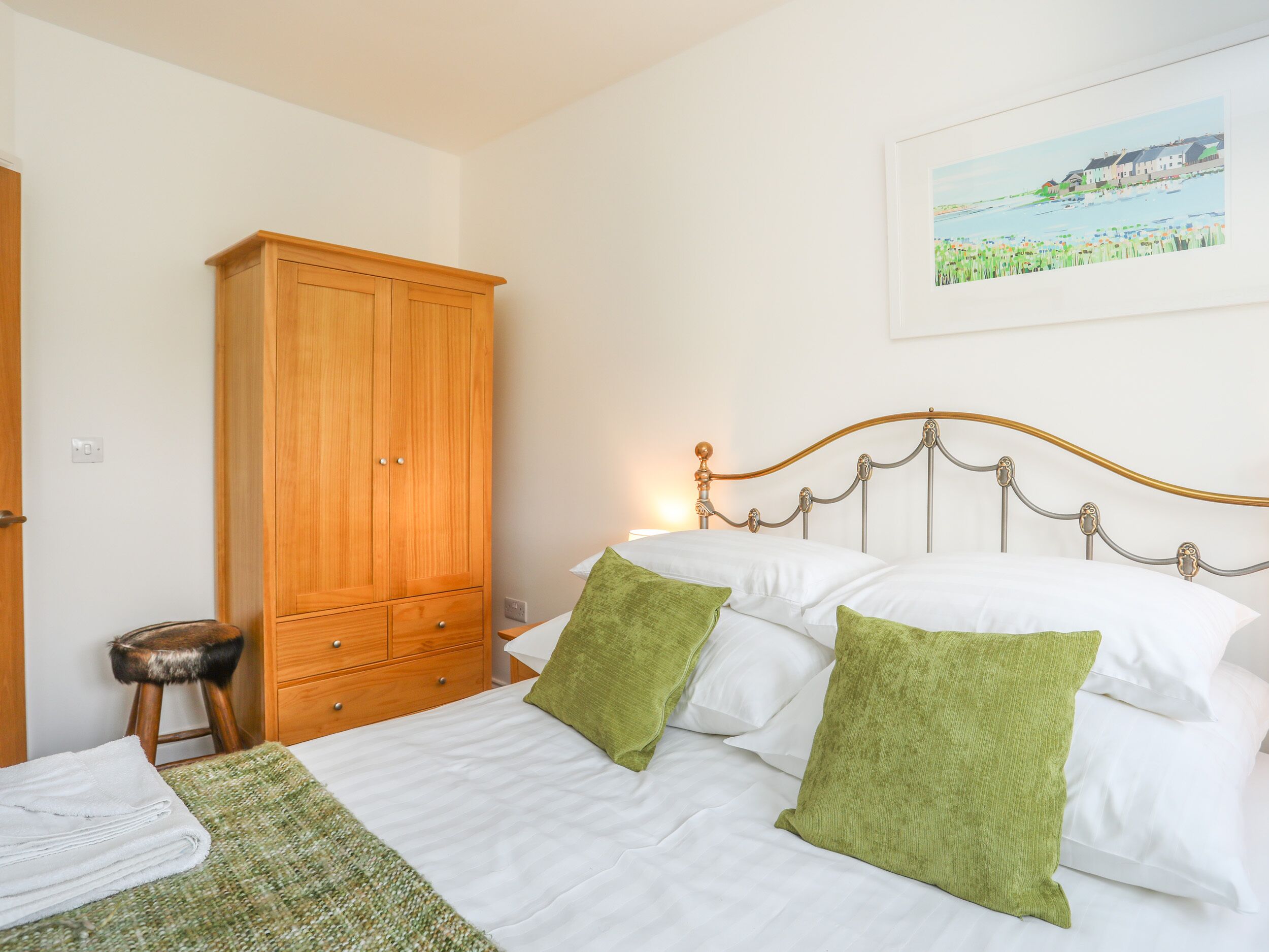 A bedroom with a bed, a wooden wardrobe, a small stool, and a painting on the wall.