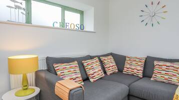 A modern living room with a grey sofa, colorful striped pillows, a wooden side table, a yellow lamp, and a wall clock with colorful hands.