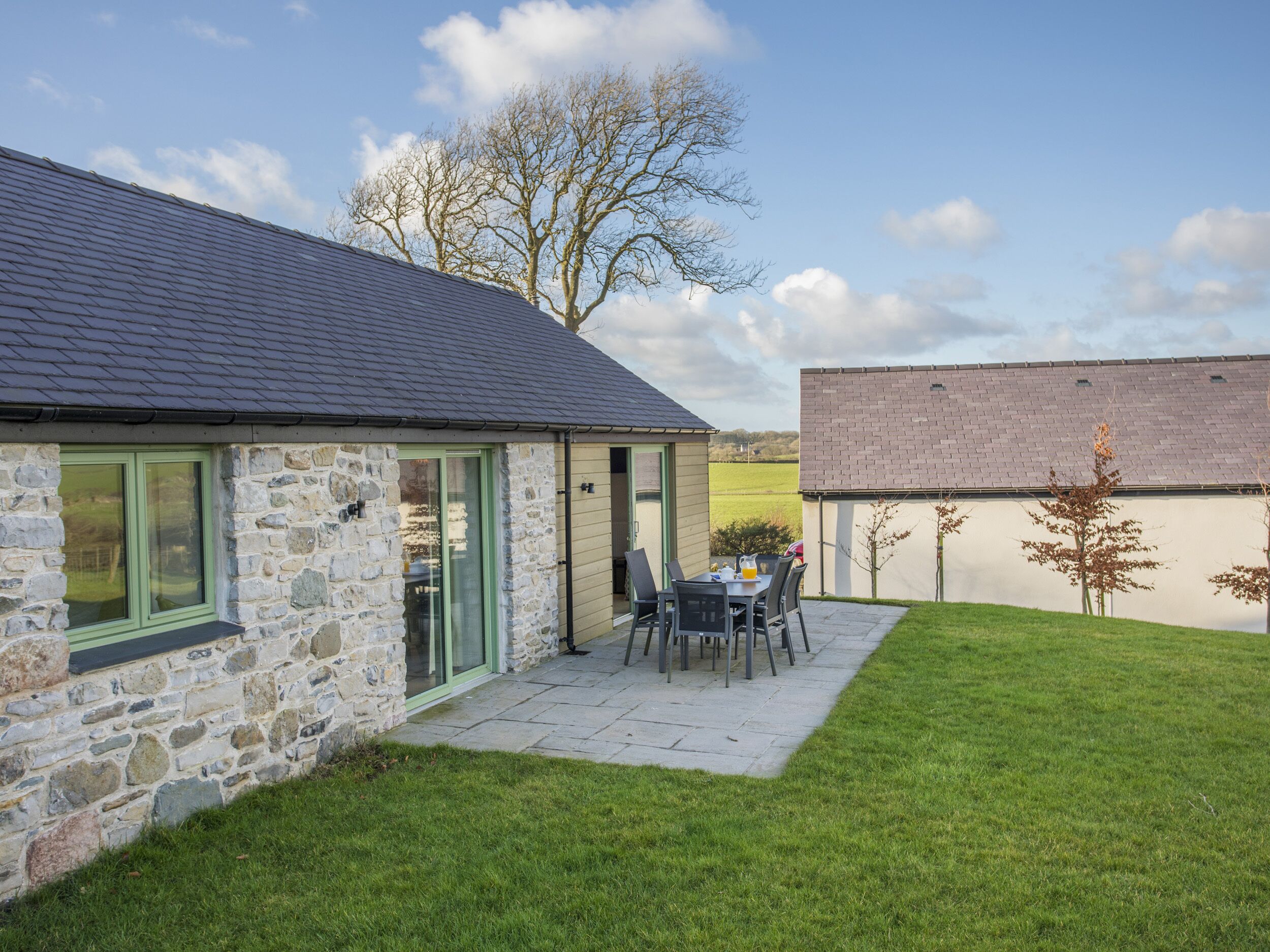 A stone building with a tiled roof, a patio with a table and chairs, and a grassy yard.