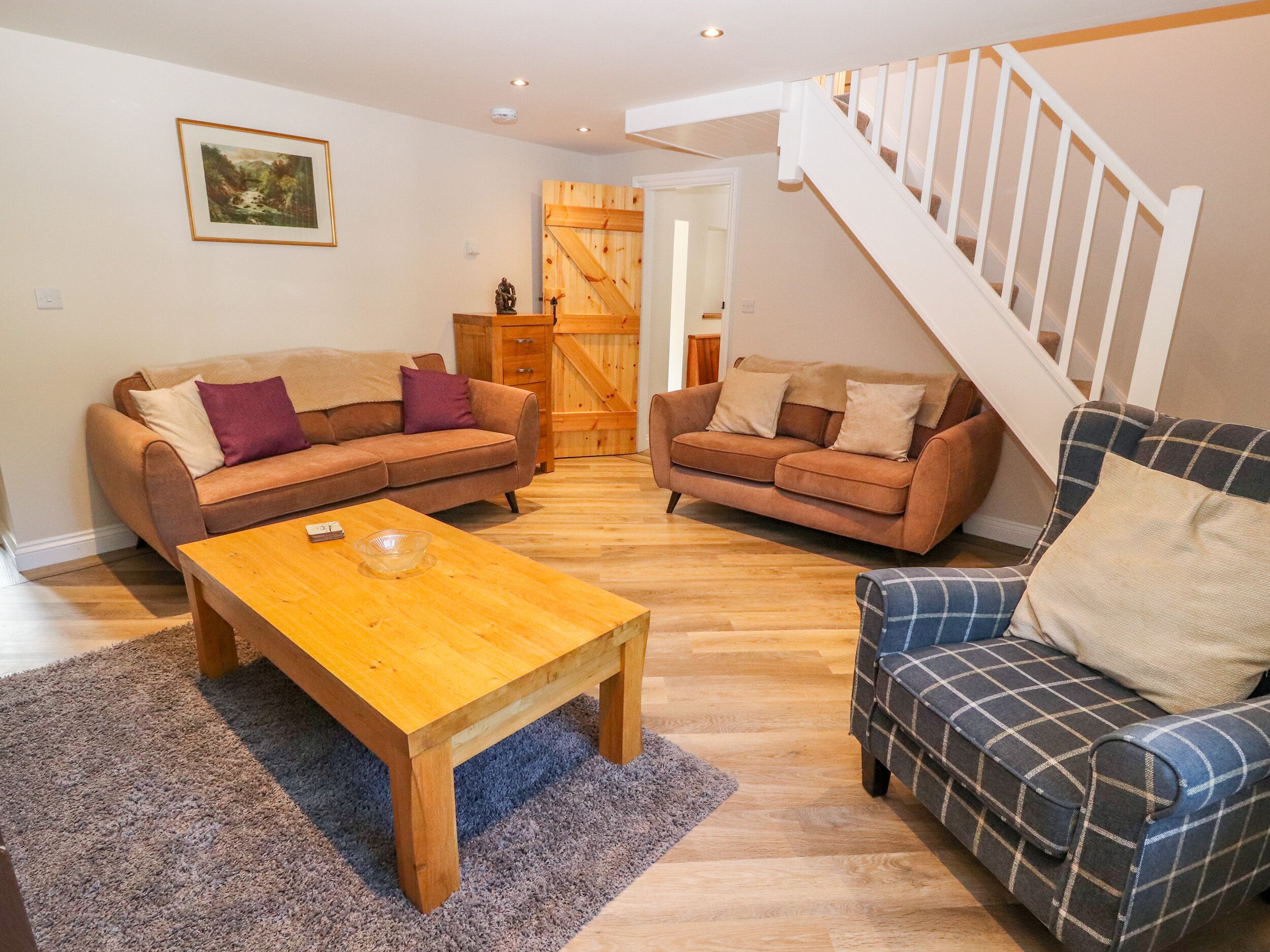 A living room with two sofas, a coffee table, and a plaid armchair. There is a staircase and a wooden door in the background.