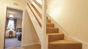 Un escalier recouvert d’une moquette beige et doté de rampes blanches mène à une chambre à coucher où se trouvent un lit et une fenêtre ornée de rideaux.