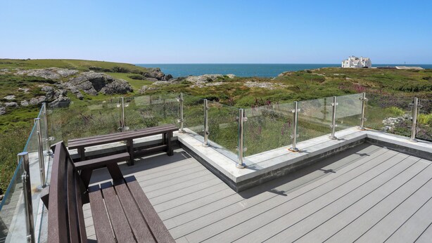 En träaltan med ett glasräcke som vetter ut mot ett kustlandskap, med ett hus synligt på avstånd.