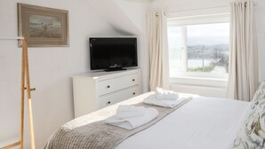A neatly arranged bedroom with a bed, a television on a dresser, a window with curtains, and a framed picture on the wall. - Ty Mawr (Menai Bridge)
