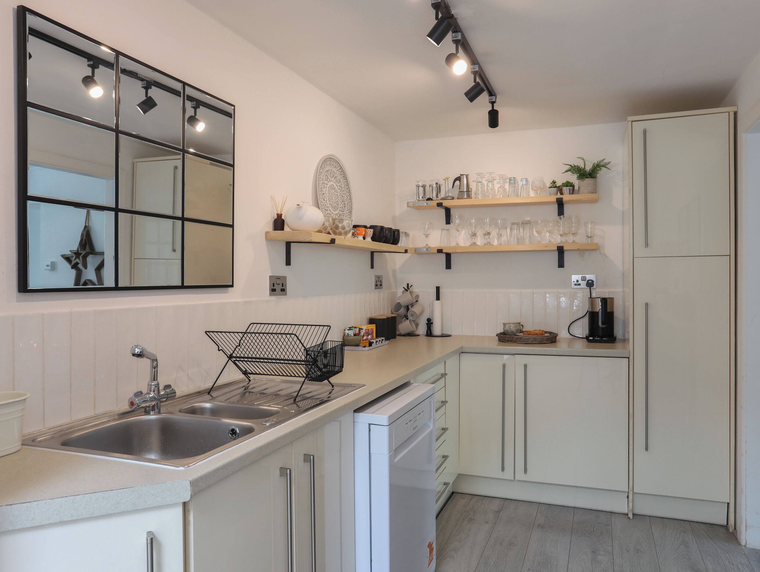 Cocina moderna con un espejo grande, pileta de acero inoxidable y muebles de cocina blancos. Se observan estantes con cristalería y una cafetera.