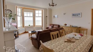 A cozy living room with a brown sofa, a dining table with a patterned tablecloth, and a view of the sea through large windows. - Beach View Apartment 3 (Weymouth)