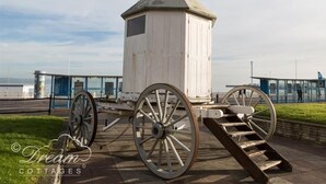A white wooden structure with a conical roof, possibly a historical building, situated on a paved area with large wheels attached to its base.