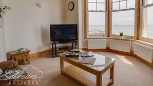 A living room with a television, a small table with a chessboard, and a large bay window with a view of the sea. - Beach View Apartment 3 (Weymouth)