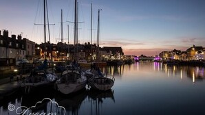 A marina with several boats docked, a calm water surface reflecting the boats and buildings, and a town with buildings along the waterfront. - Beach View Apartment 3 (Weymouth)