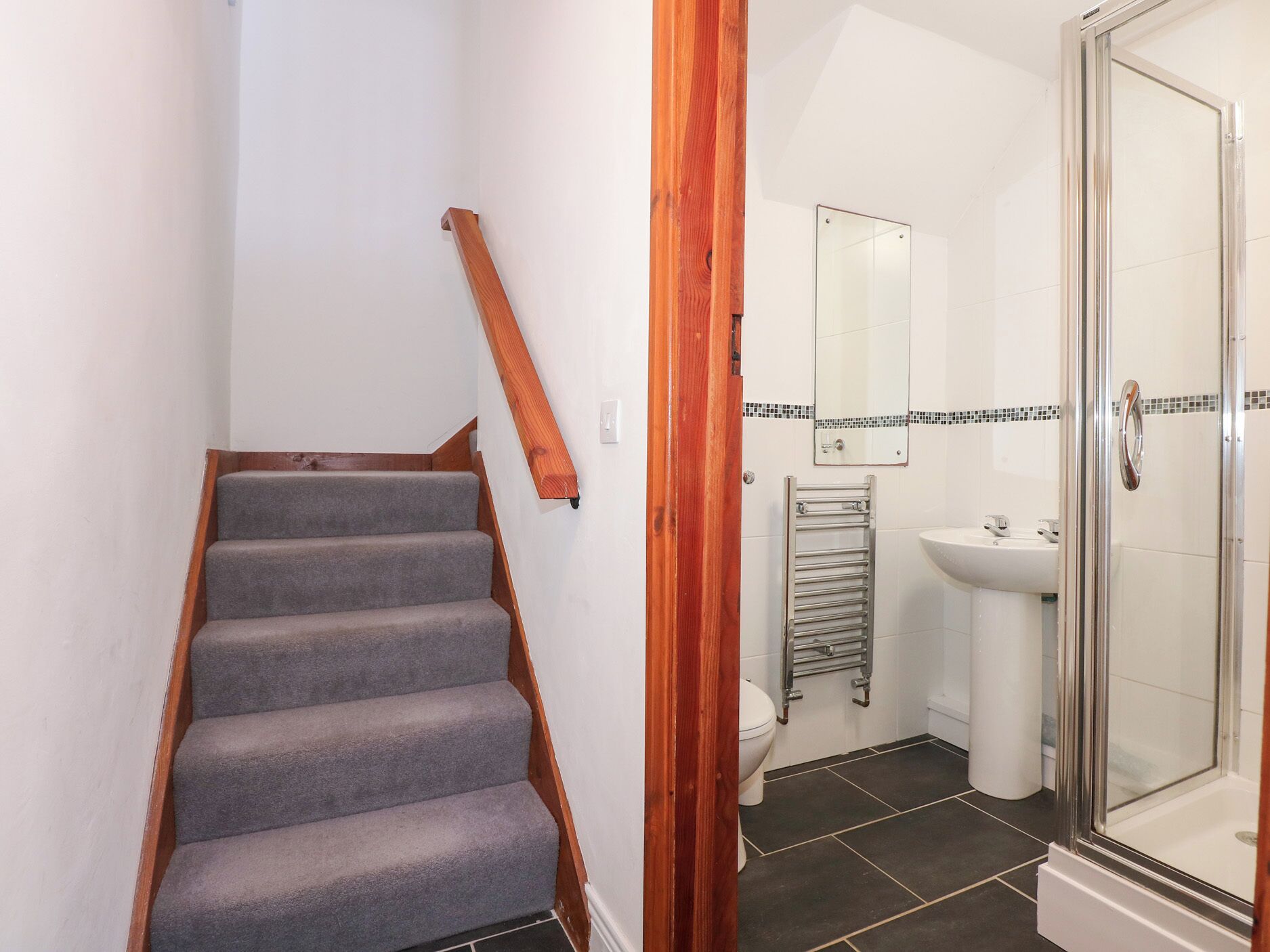 Un escalier avec des marches recouvertes de moquette grise et des rampes en bois, menant à une salle de bain équipée d’une douche, d’un lavabo et de toilettes.
