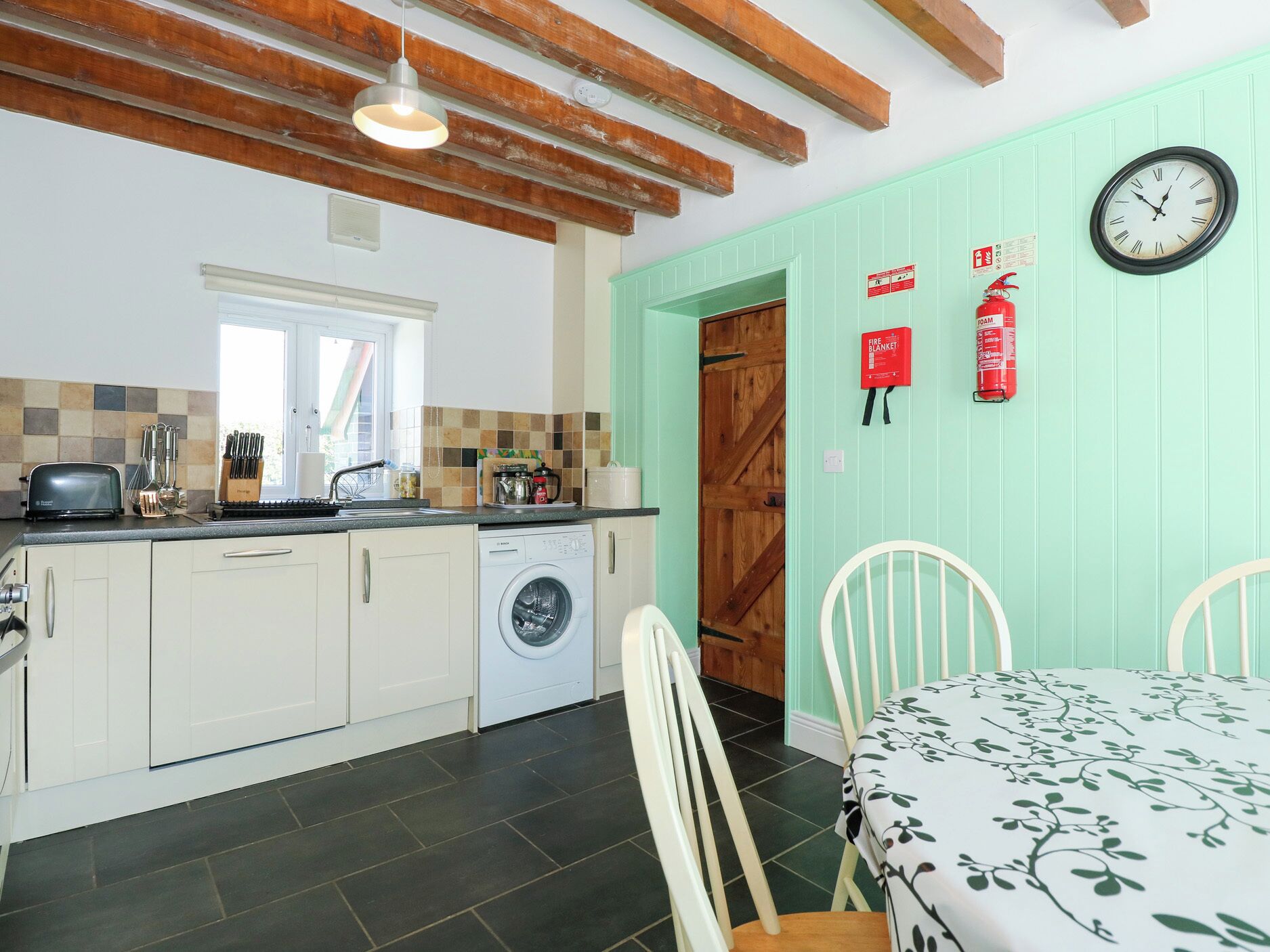 Een keuken met een wasmachine, een eettafel met stoelen, een klok en een veiligheidssignaal voor brand.