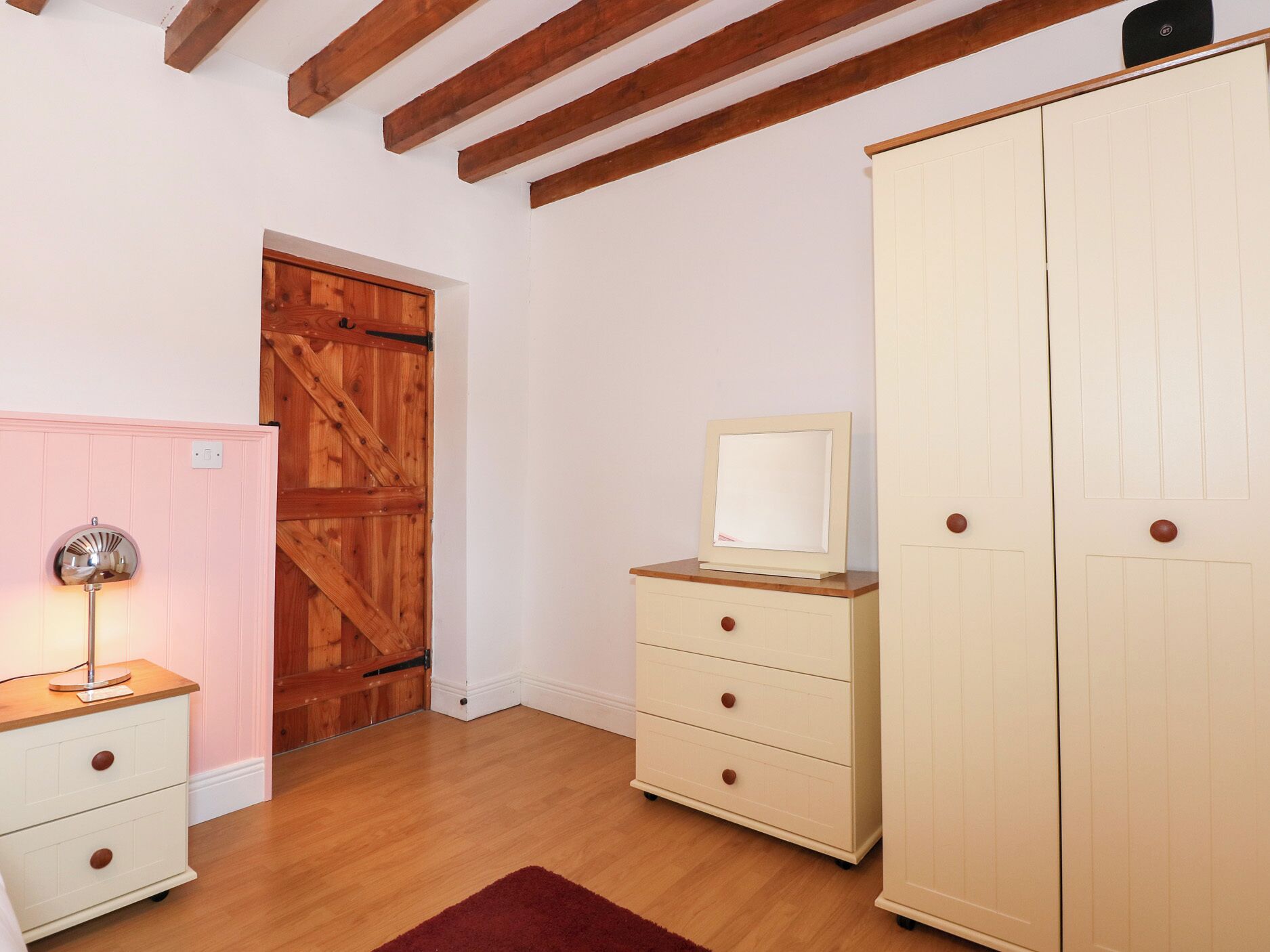 Una habitación con vigas de madera en el techo, una puerta de madera, un aparador blanco con cajones, un espejo encima y un armario grande.
