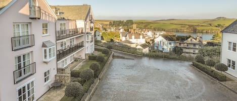 A courtyard with a cobblestone path, surrounded by buildings with balconies and a view of a village and water body in the distance.