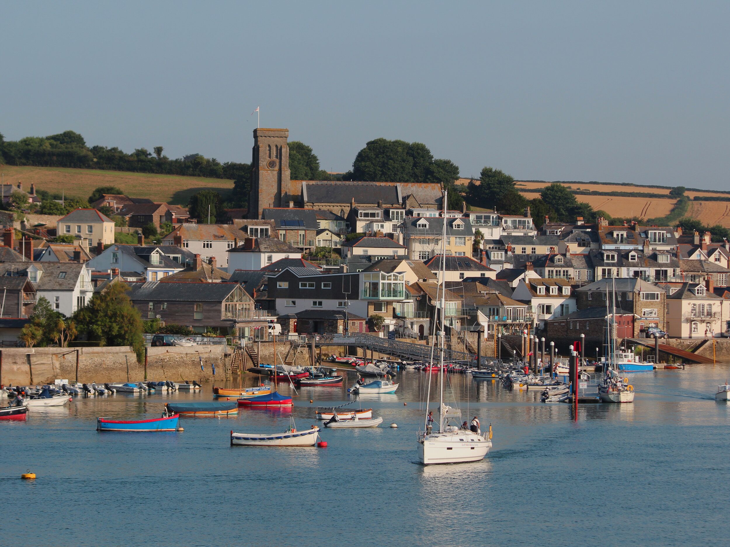 Une ville côtière dotée d’une tour imposante, d’un port de plaisance où sont amarrés différents types de bateaux, et d’un quartier résidentiel densément peuplé, composé de maisons.