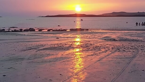 A beach at sunset with a clear sky, calm water, and distant mountains. - West View (Rhosneigr)