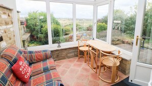 A conservatory with a wicker table and chairs, a plaid sofa with a cushion, and a view of the garden.