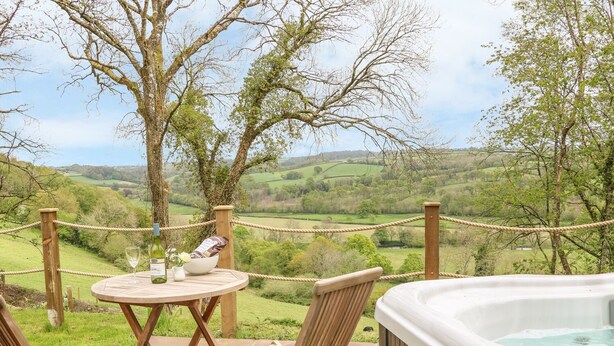 A hot tub on a patio with a table and two chairs, overlooking a scenic countryside.