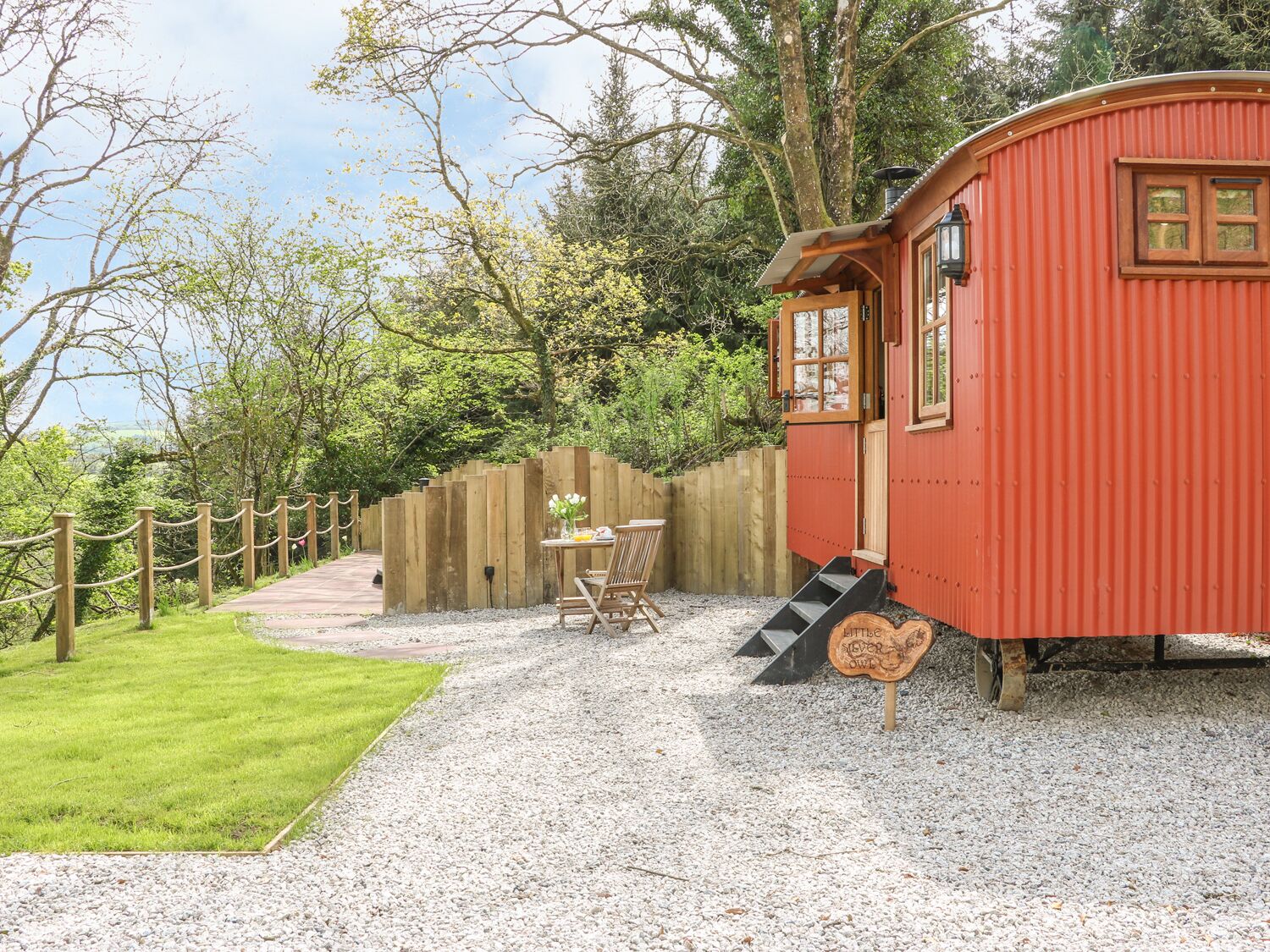 A red wooden cabin with a gravel path leading to it, surrounded by greenery and a wooden fence.