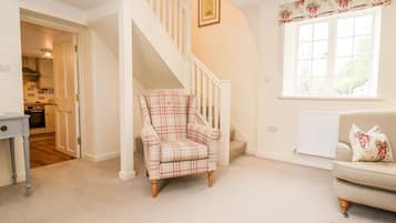 A living room with a staircase, a sofa, a plaid armchair, and a window with floral curtains.