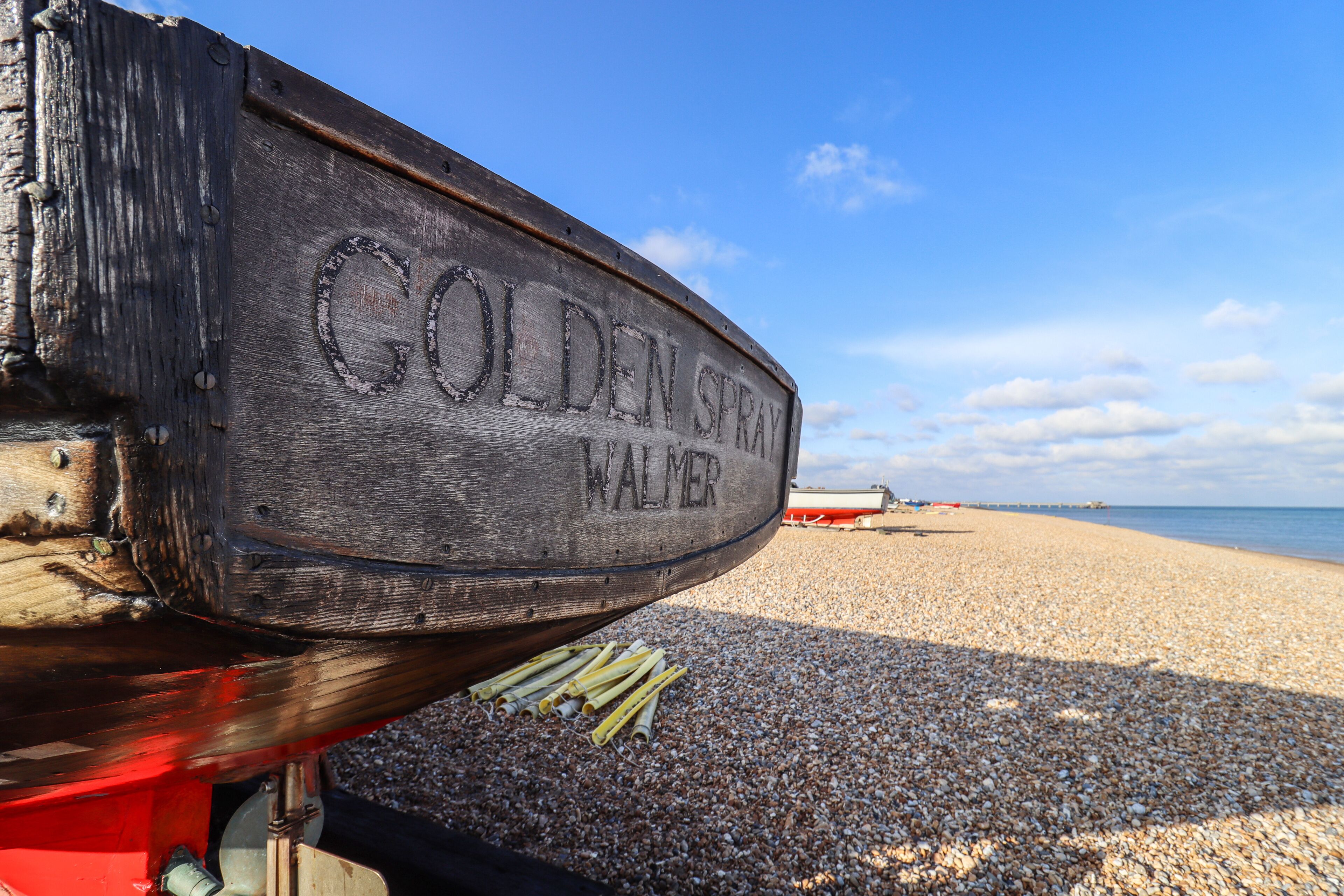 Una señal de madera con la inscripción "GOLDEN SPRAY WALMER" en una playa de guijarros.