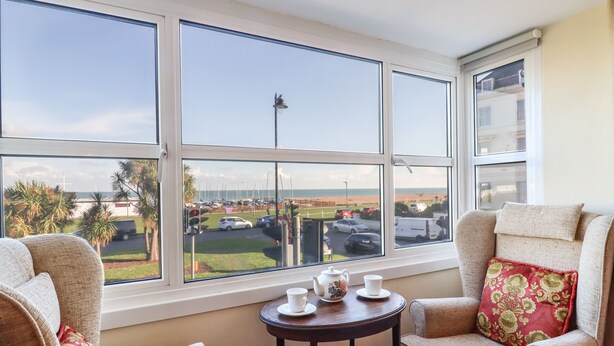 Ein Zimmer mit großem Fenster mit Blick auf eine Marina und einen Strand, ausgestattet mit zwei Sesseln und einem kleinen Tisch mit Teekanne und Tassen.