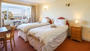 Ein Hotelzimmer mit zwei Betten, einem Schreibtisch, einem Stuhl, einem kleinen Tisch und Meerblick.