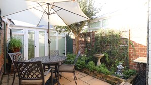 Eine Terrasse mit Tisch und Stühlen unter einem großen Sonnenschirm, umgeben von einer Ziegelmauer und Grünpflanzen.