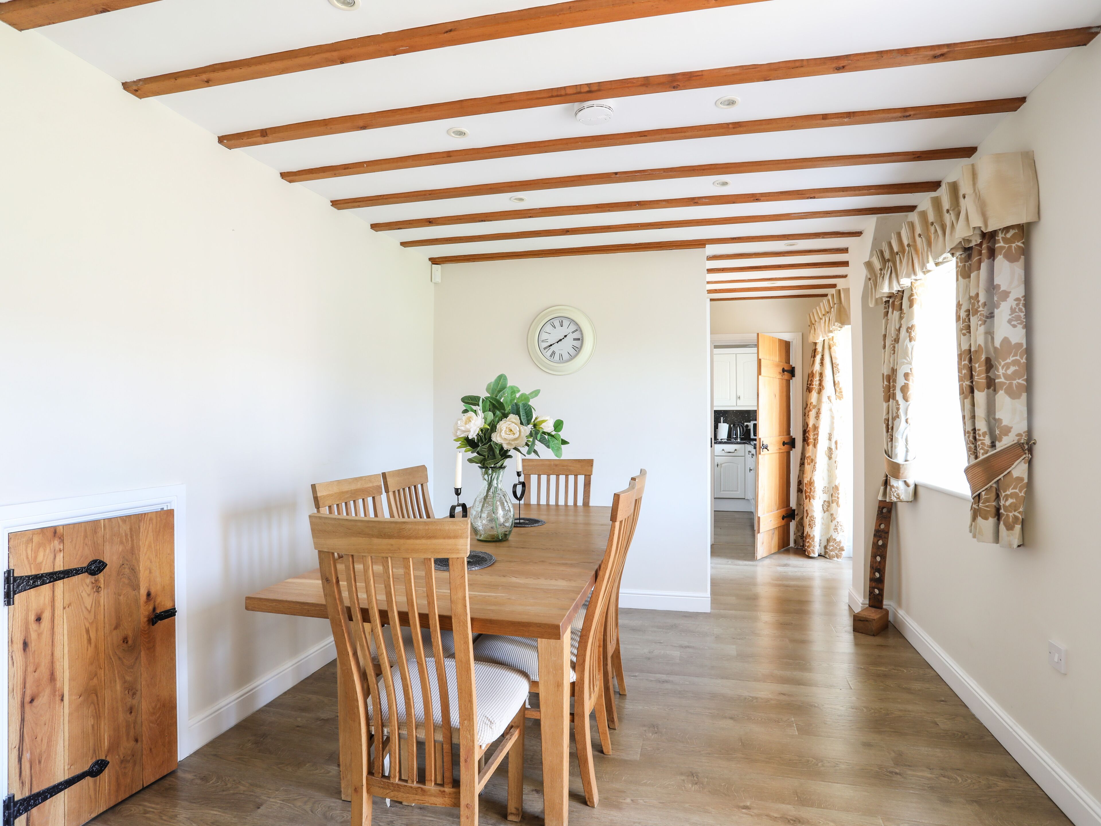Un comedor con sillas de madera y una mesa, un reloj en la pared y una cocina al fondo.