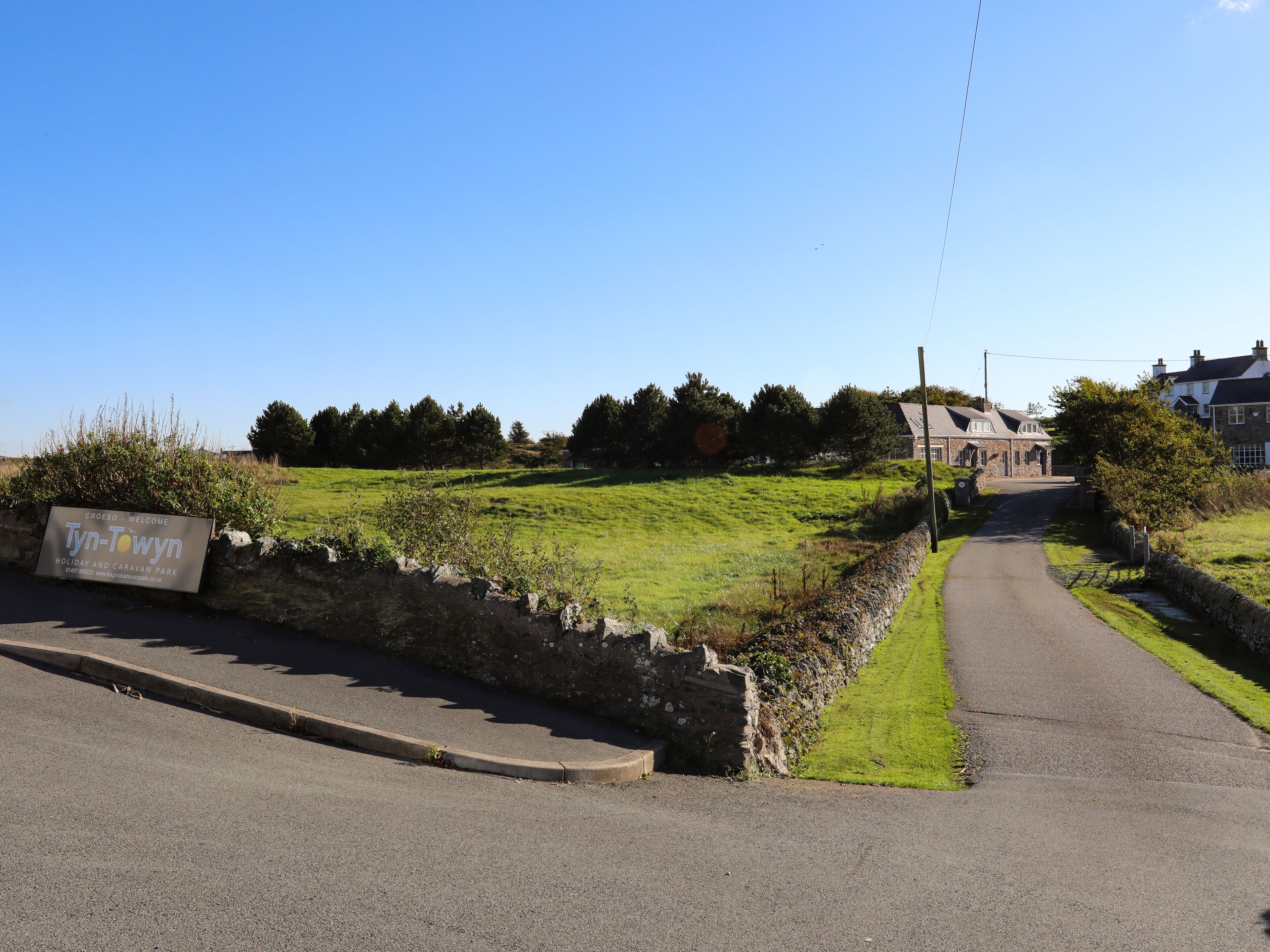 A rural road leading to a stone house with a grassy field and trees in the background.