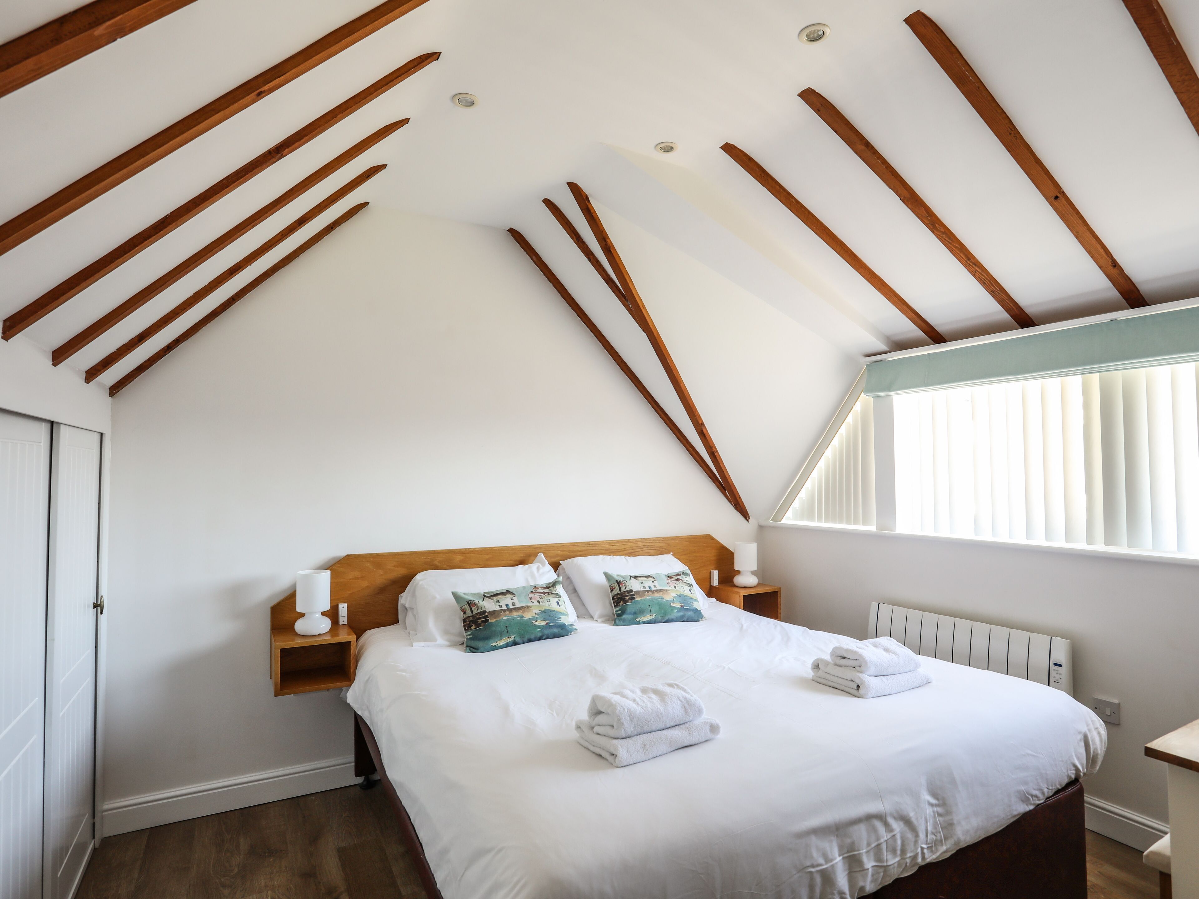 Une chambre avec un plafond en pente, des poutres en bois, un lit avec du linge de lit blanc, deux coussins décoratifs, une table de chevet avec une lampe et une fenêtre avec des stores.
