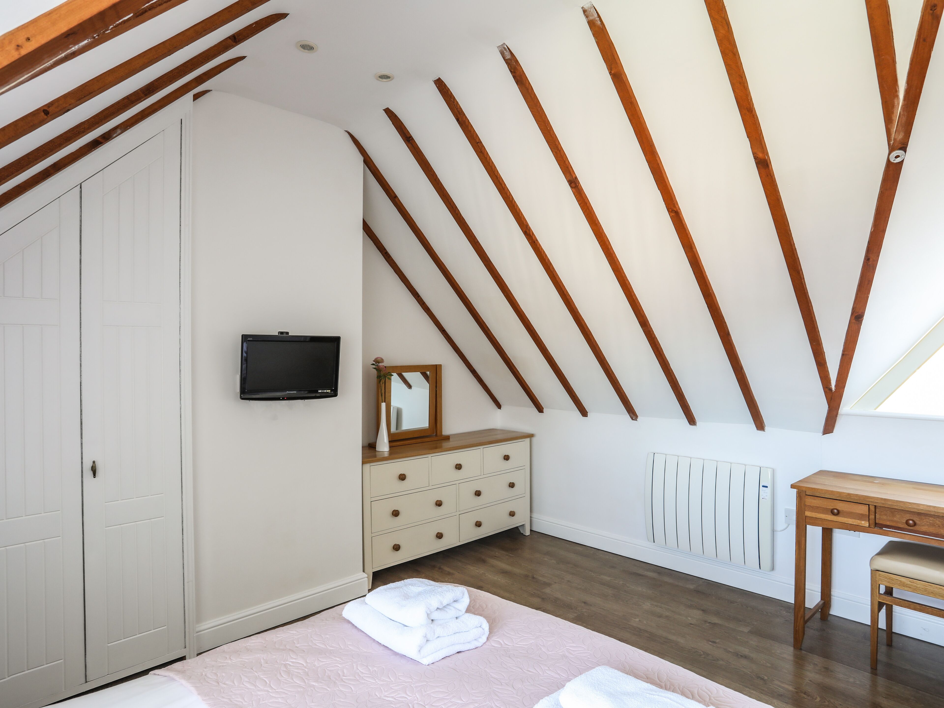 Een slaapkamer met schuine wanden, houten balken, een witte garderobe, een dressoir, een spiegel, een televisie, een radiator, een houten bureau en een stoel.