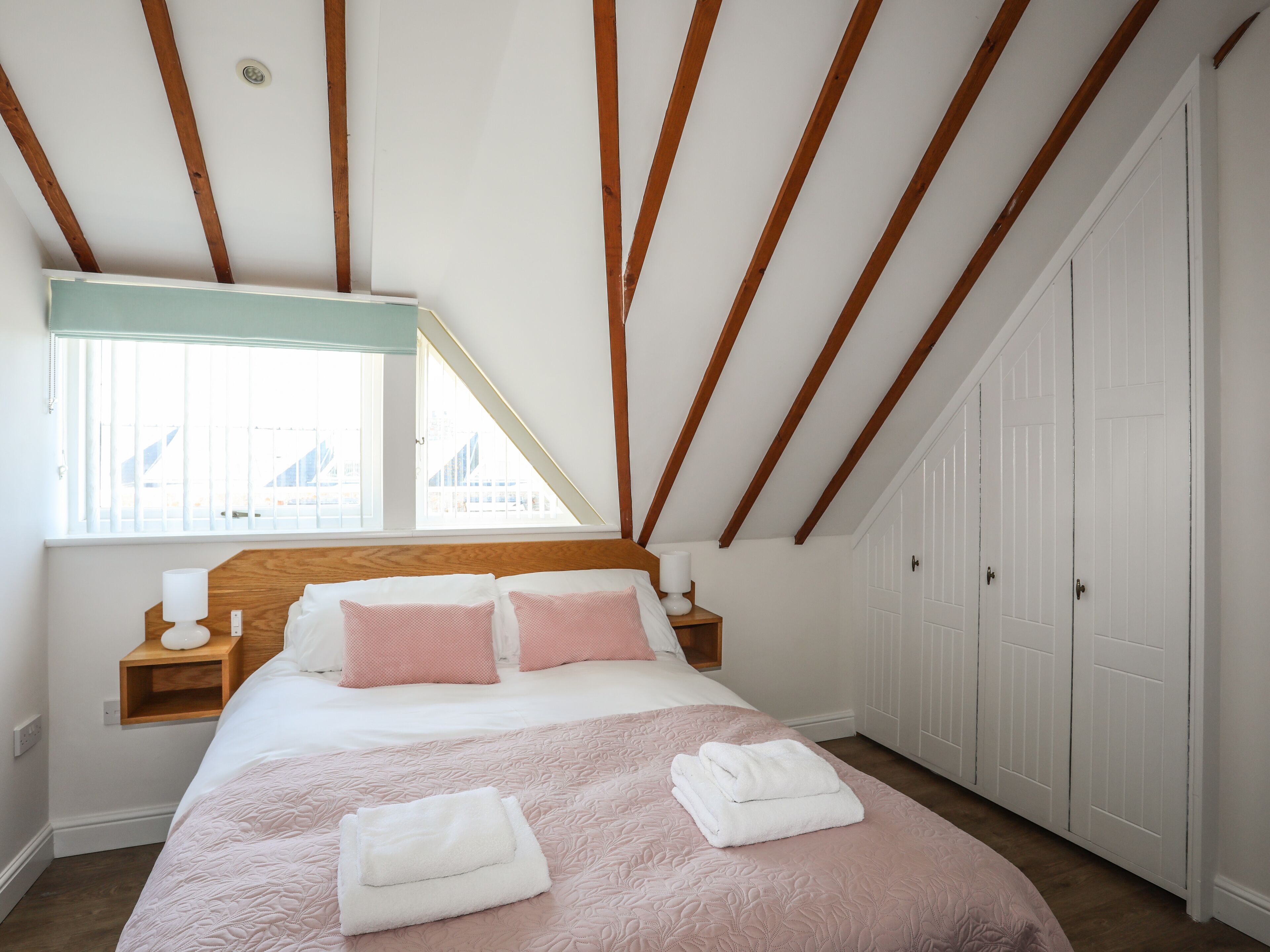 Un dormitorio con techo inclinado, una cama con ropa de cama rosa, dos mesitas de noche con lámparas y una ventana con vistas a edificios.