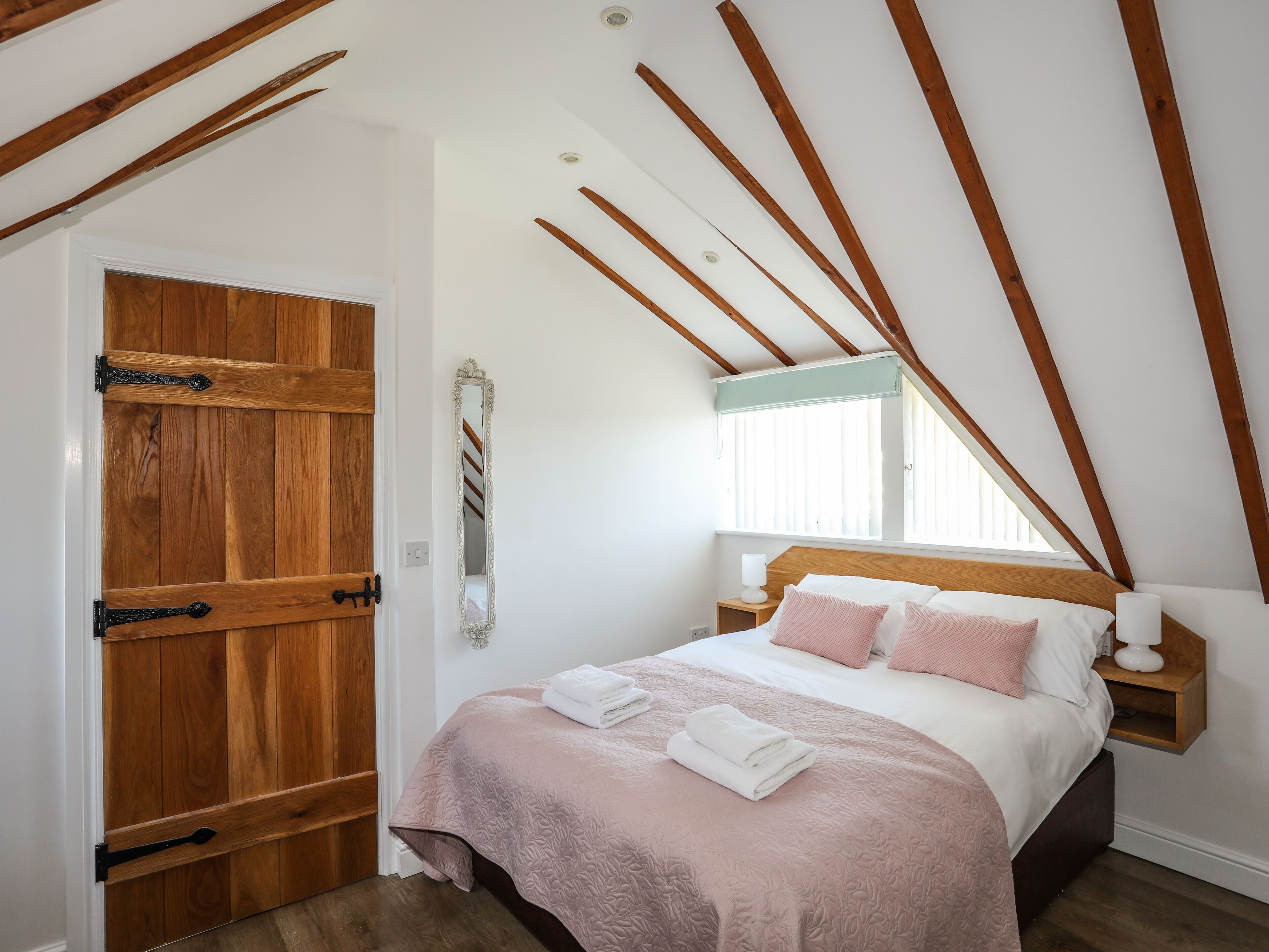 A bedroom with a sloped ceiling, wooden beams, a bed with pink bedding, a bedside table with a lamp, and a wooden door.
