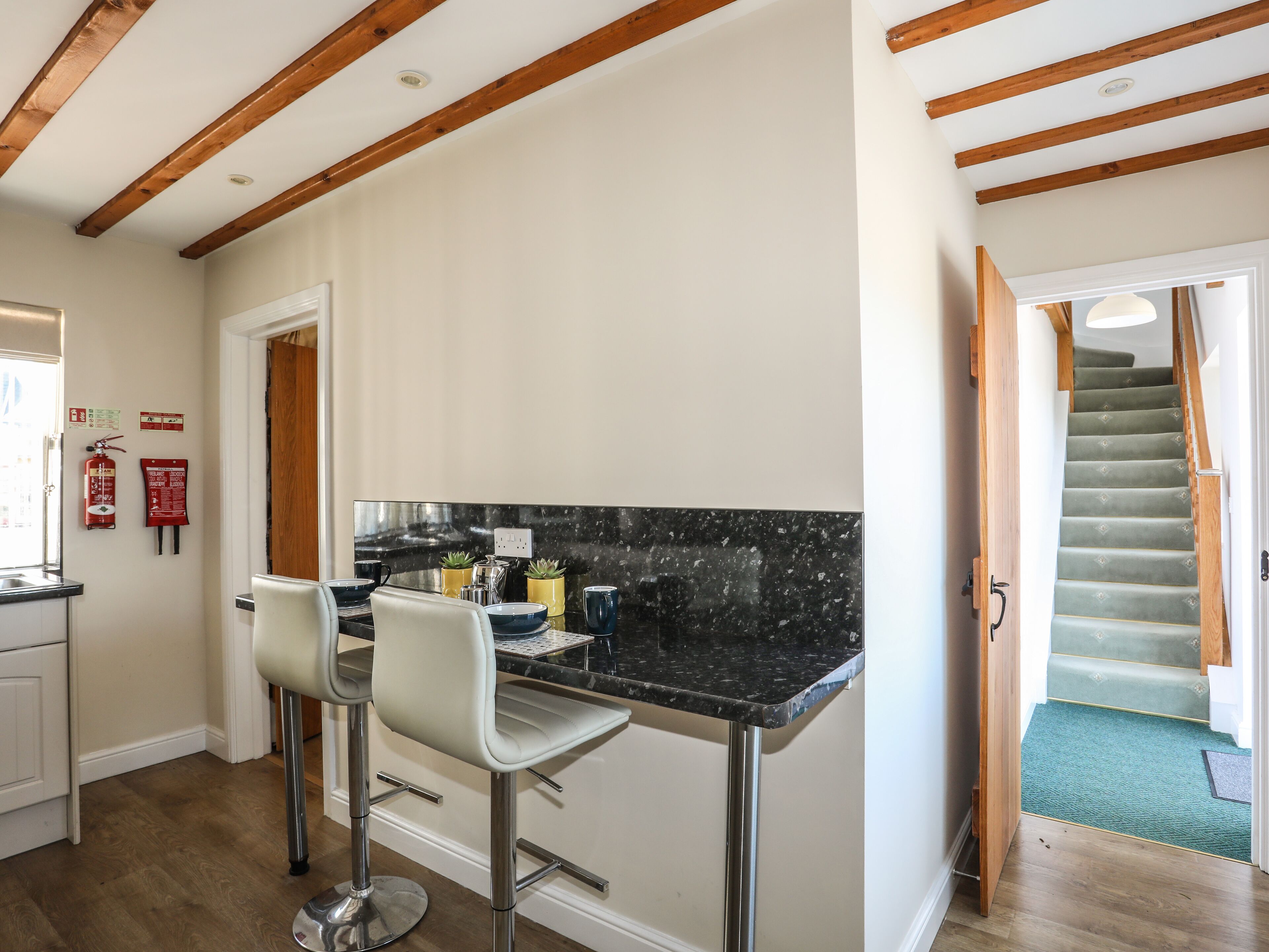 A kitchen with a black marble countertop, two white bar stools, wooden beams on the ceiling, and a staircase leading to a landing.