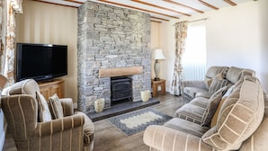 A living room with a stone fireplace, wooden beams, a flat-screen TV, and striped sofas. - Tyn Towyn - Ty Gwyrdd (Holyhead)