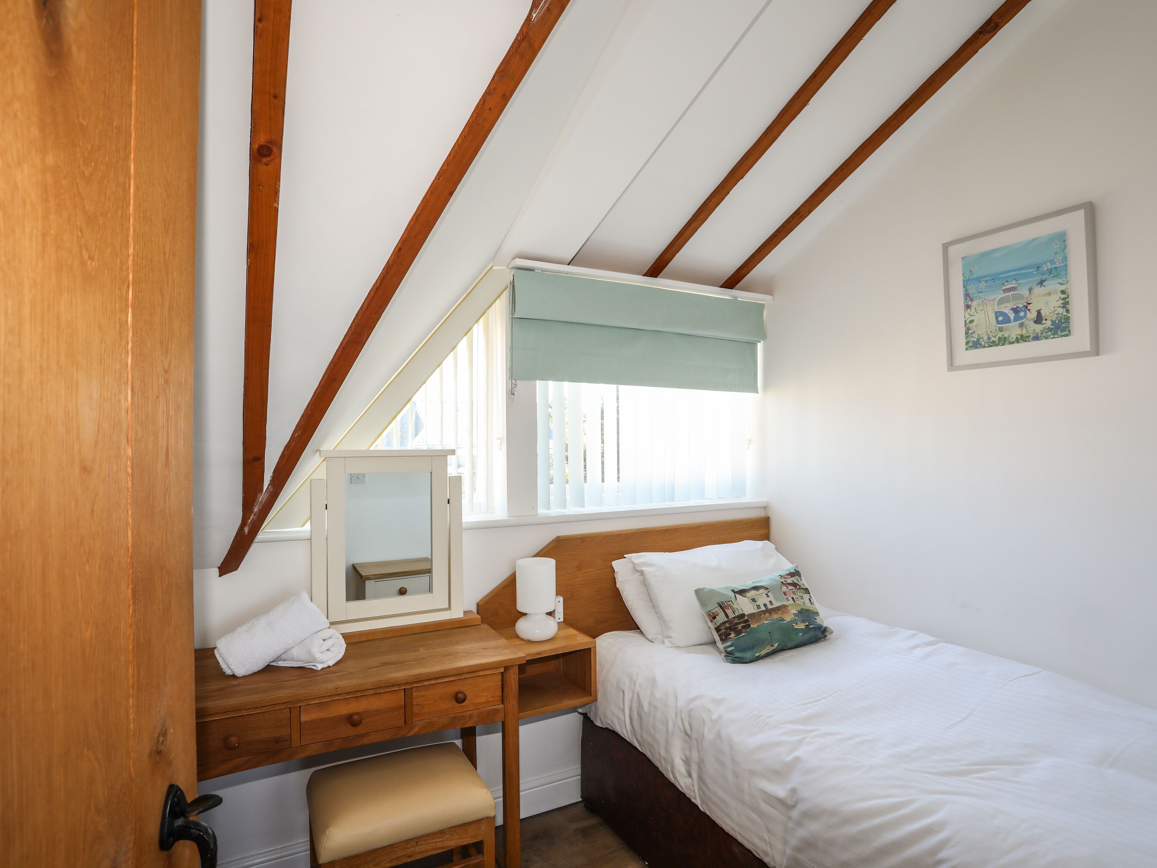 A bedroom with a wooden desk, a bed with white bedding, a window with a view, and a painting on the wall.