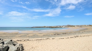 A beach with a clear sky, rocky foreground, and a distant town. - Tyn Towyn - Ty Oren (Holyhead)
