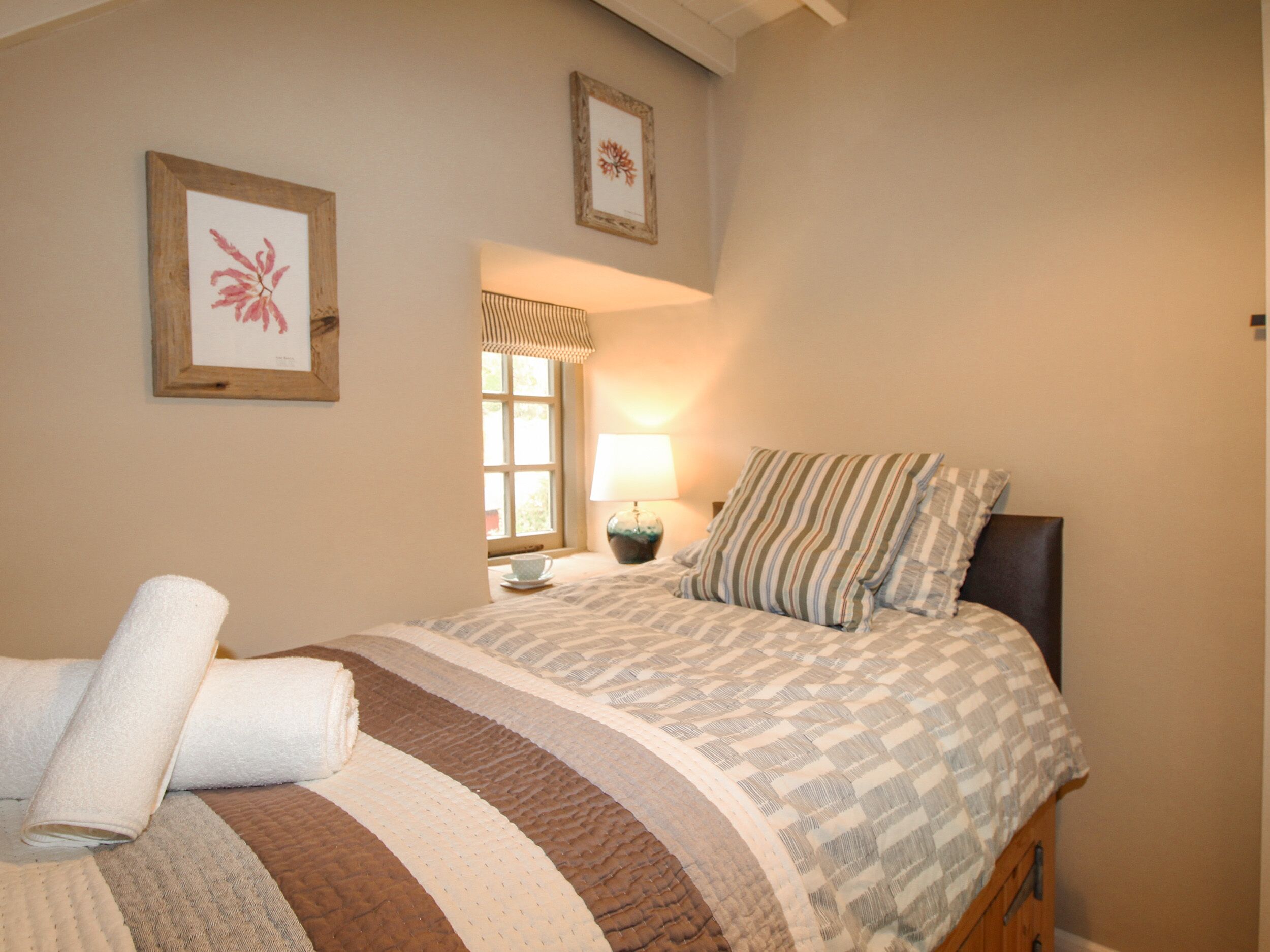 Ein Schlafzimmer mit Bett, Fenster, Lampe und zwei gerahmten Bildern an der Wand.