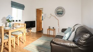 A modern living room with a wooden dining table, a leather sofa, a TV, and a decorative wall piece. - Tyn Towyn - Bwthyn Haf (Holyhead)