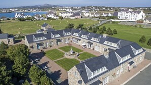 A large stone building with multiple wings, a central courtyard with a fountain, and a view of a coastal town in the background. - Tyn Towyn - Bwthyn Haf (Holyhead)