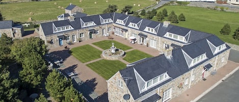 A large stone building with multiple wings, a central courtyard with a fountain, and a view of a coastal town in the background.