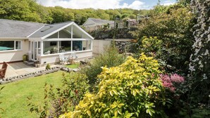 A modern house with a large glass conservatory, a stone wall, and a garden with various plants and flowers. - Clyd Fan (Harlech)