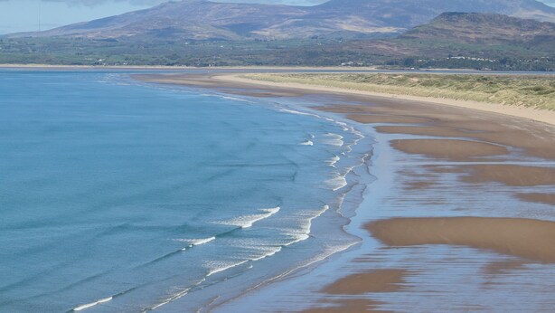 A coastal landscape with a sandy beach, gentle waves, and distant mountains under a clear sky. - Clyd Fan (Harlech)