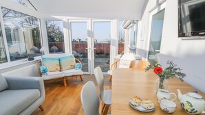 A sunlit room with a wooden dining table, a grey sofa, and a large window offering a view of the outside. - Clyd Fan (Harlech)